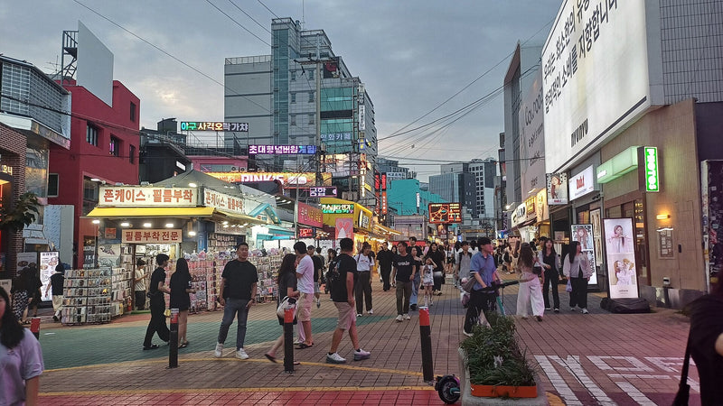 Hongdae shopping street at dusk, lined with photo booths and idol-poster shops where young K-pop fans gather