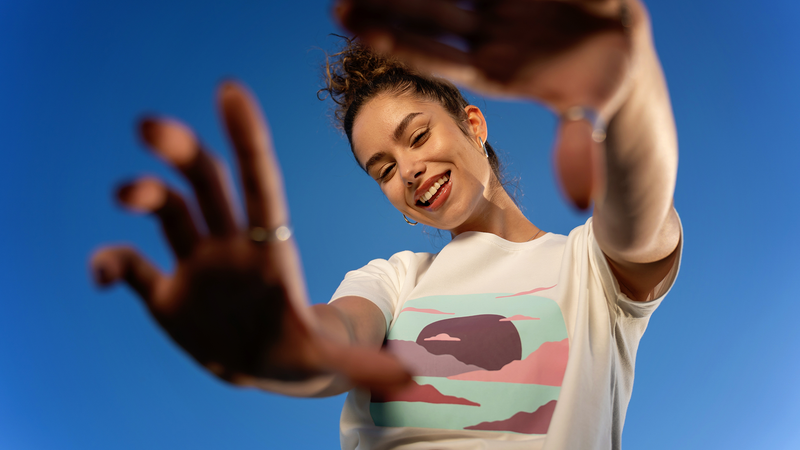 グラフィックTシャツ姿の若い女性が、明るい青空を背にカメラへ手を伸ばしている