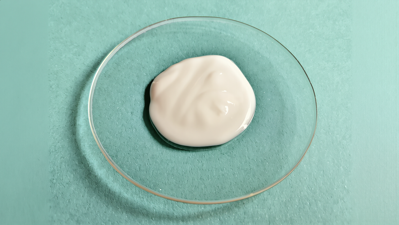 A dollop of white moisturizing cream on a clear glass dish, set on pale celadon paper in soft morning light