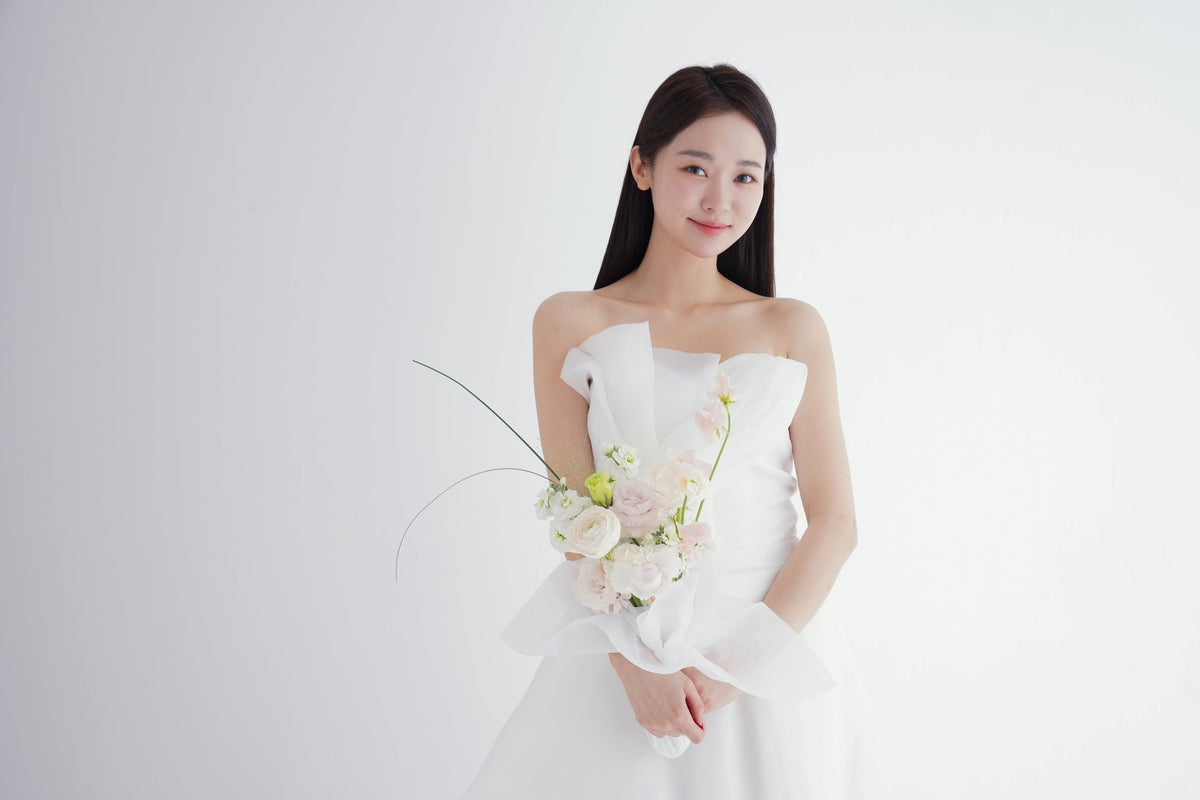 白い壁の前で淡いピンクのブーケを持つ、ストラップレスの白いドレス姿の花嫁