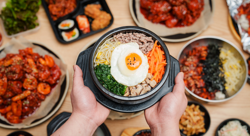 目玉焼きをのせた熱々の石焼ビビンバを手に持ち、周りに韓国の小皿料理が並ぶ様子