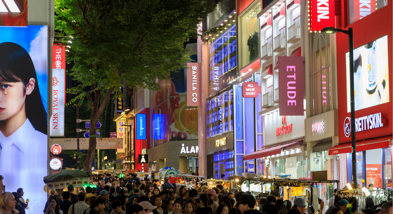 Myeongdong's main street at night, crowds passing between brightly lit K-beauty storefronts