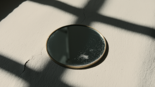 A small round hand mirror on pale plaster with the hard shadow of a window frame falling across it