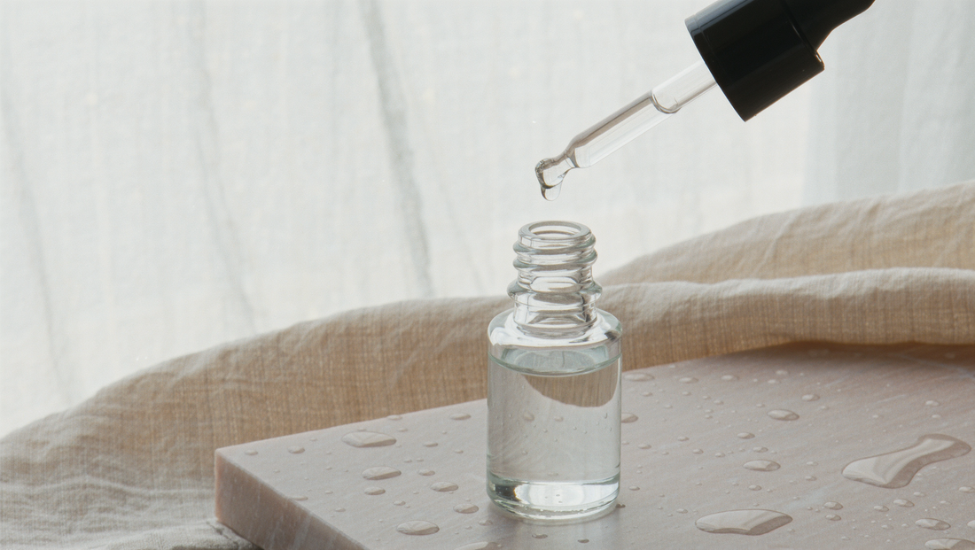 A glass dropper bottle with a pipette hovering above it on pale limestone and linen in soft morning light