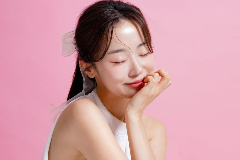 ピンクの背景の前で、ほおづえをついて微笑むツヤ肌の女性