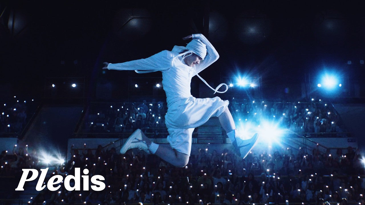 A TWS member leaps above a sea of glowing lightsticks on a concert stage in the Being Together : TO US video