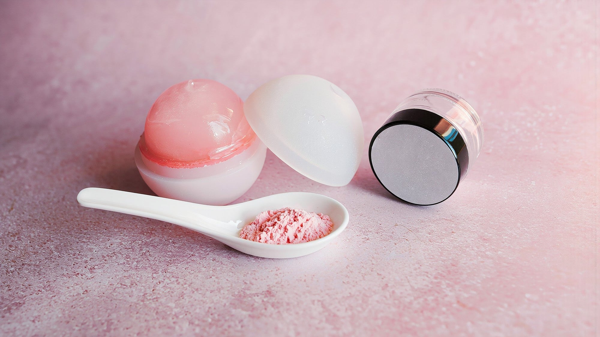 PDRN serum still life: pale pink skincare jars and a spoon of powder on a soft pink surface in bright morning light
