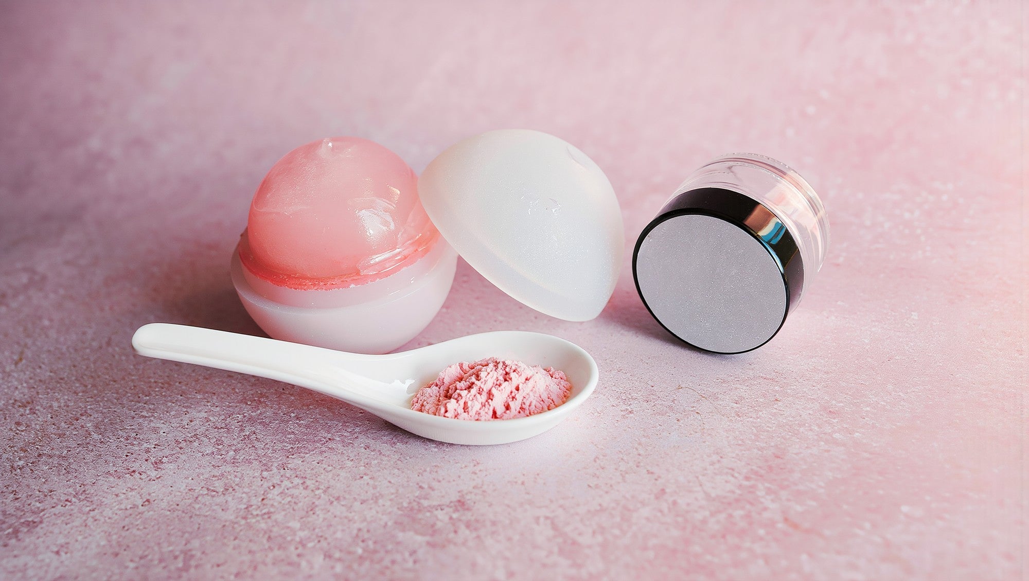 PDRN serum still life: pale pink skincare jars and a spoon of powder on a soft pink surface in bright morning light