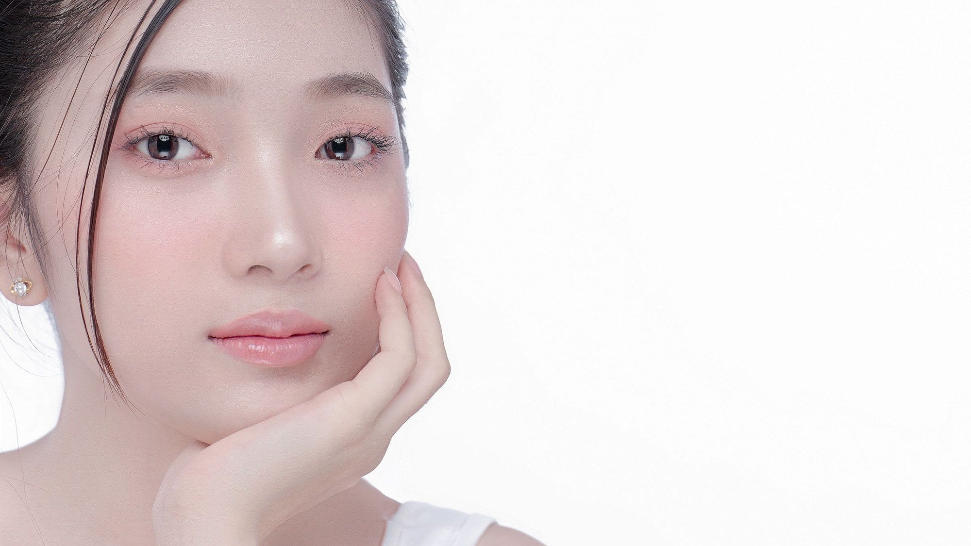 Skin-first beauty look: a young woman with dewy, lightly made-up skin rests her chin on her hand against a white backdrop