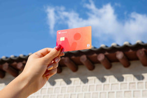 WOWPASS prepaid travel card, which carries a T-money transit chip, held up against a hanok tiled roof and blue sky (WOWPASS)